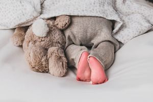 A plush teddy bear sits beside pink bunny slippers on white bedding.