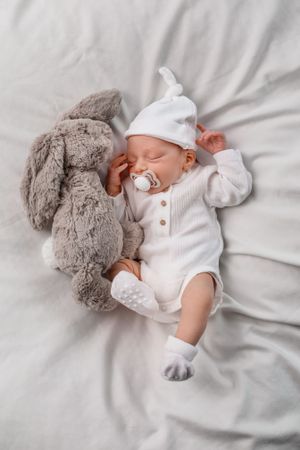 Sleeping newborn in white outfit cuddling with plush grey bunny on soft bedding.