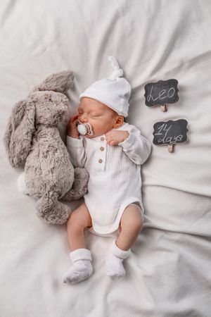 Sleeping baby with white hat and grey bunny toy next to small chalkboard signs.