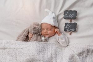 Peaceful sleeping newborn with white hat and grey bunny beside chalkboard signs.