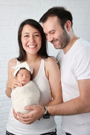 A newborn baby wrapped in beige swaddling cloth is held close to the camera against a white backdrop.