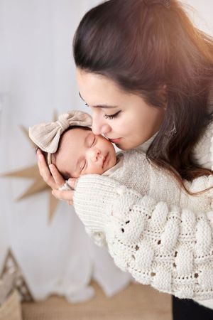 A sleeping newborn with a beige bow is tenderly held against a white backdrop.