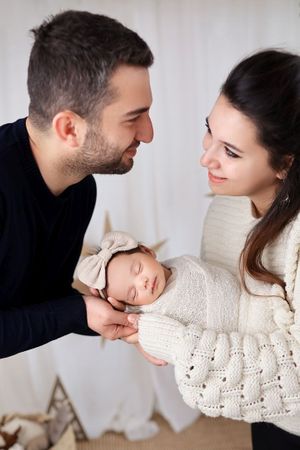 A sleeping newborn in beige swaddle and bow is held lovingly against a bright background.