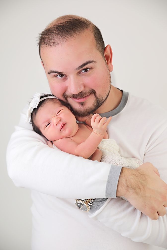 Sweet moment between parent and sleeping newborn in white outfits against light background.