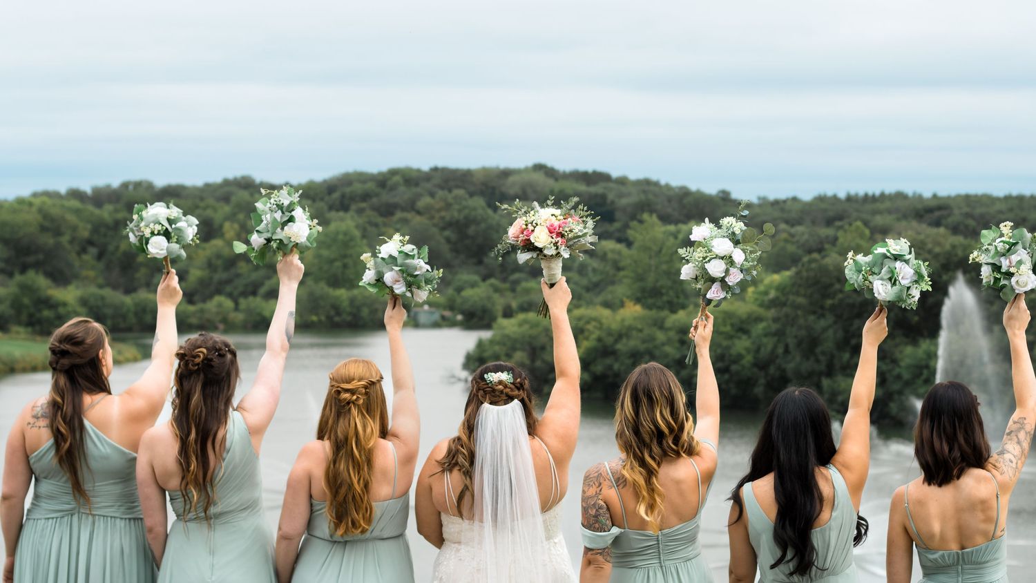 Bride and bridesmaids facing towards lake and trees in Lake Geneva Wisconsin on wedding day with bouquets in the air.