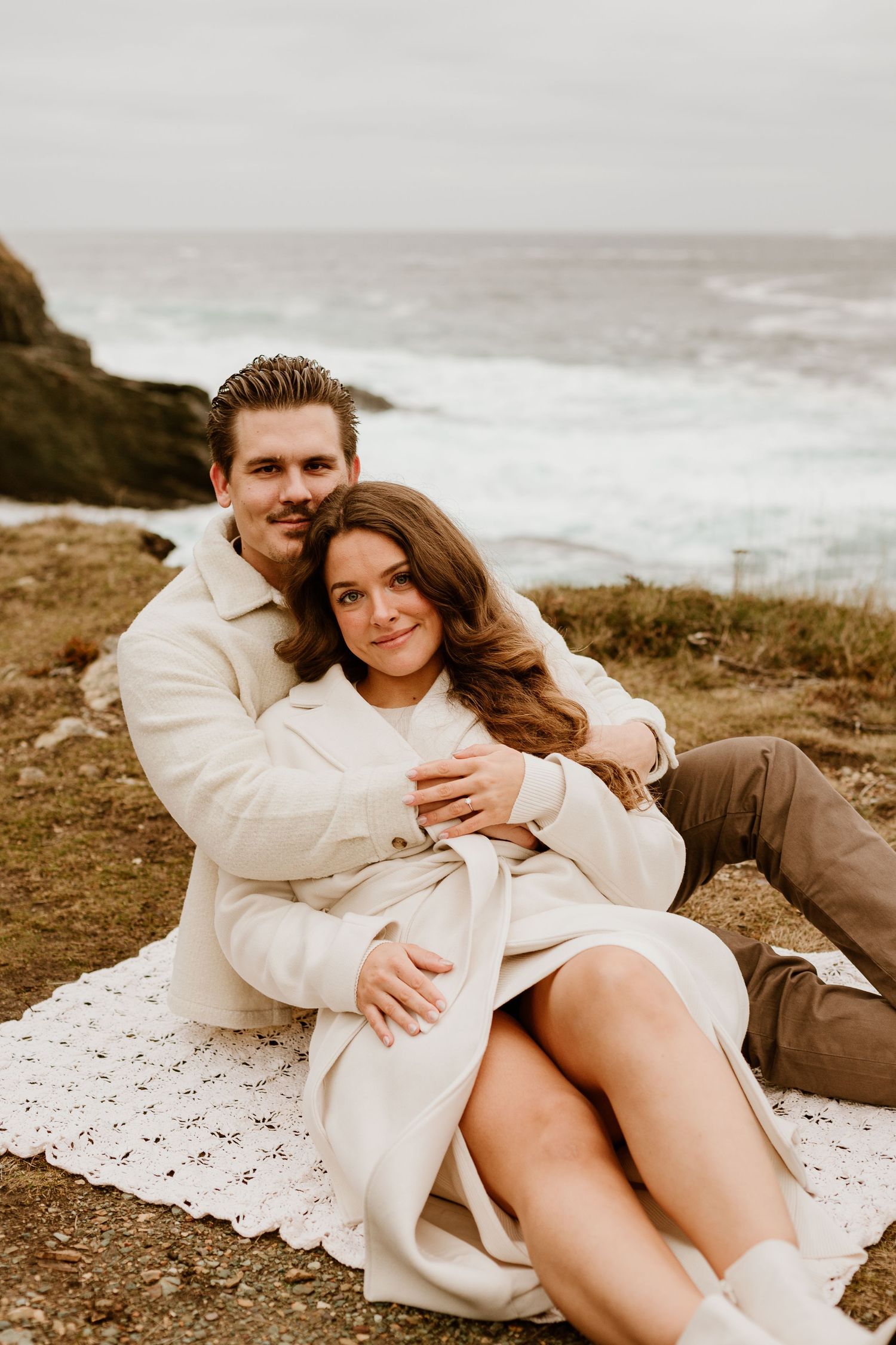 engaged couple sitting on a lace blanket on the Newfoundland beach, cuddling and smiling at the camera