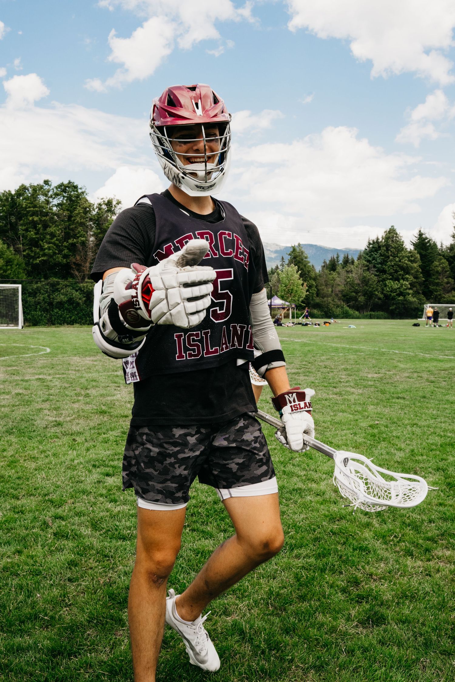 Lacrosse player in Rhode Island gear poses with stick on sunny grass field with mountains in background.