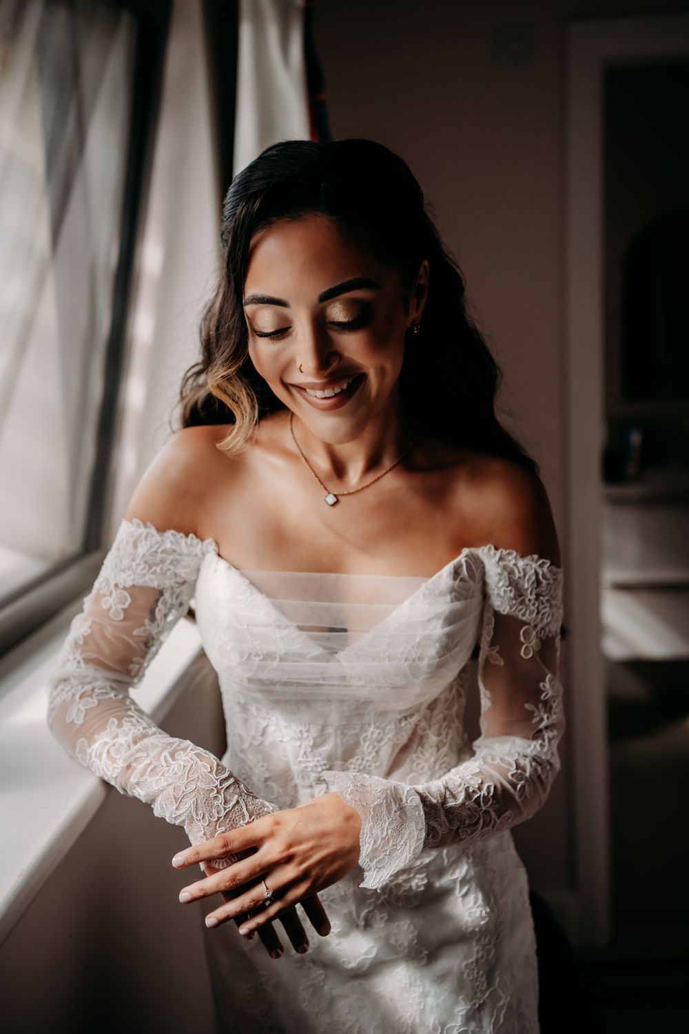 A bride in an elegant lace wedding dress with off-shoulder sleeves poses near a window in dramatic lighting.