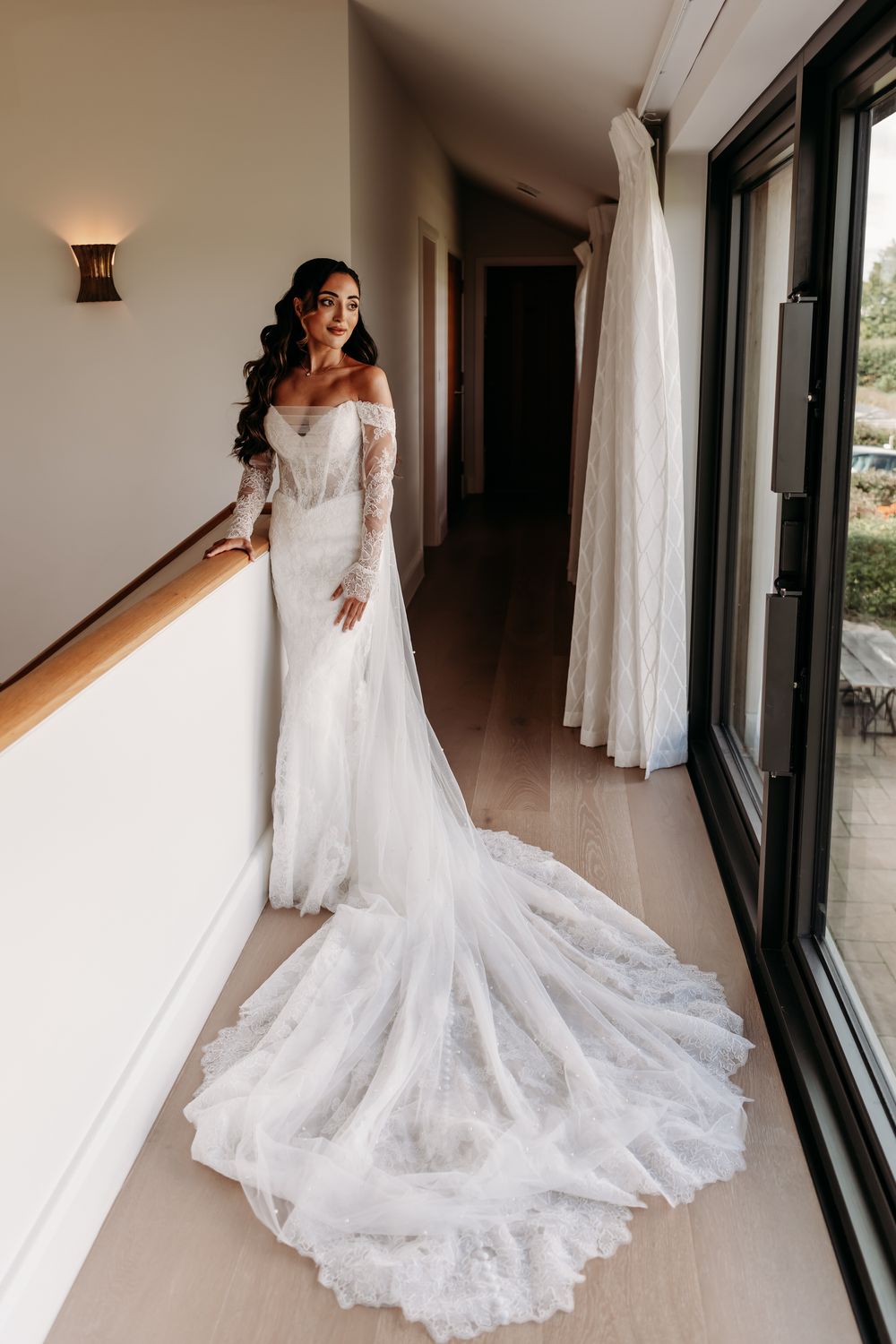 A bride in a flowing white lace wedding gown with a dramatic train poses in front of large windows in a hallway.