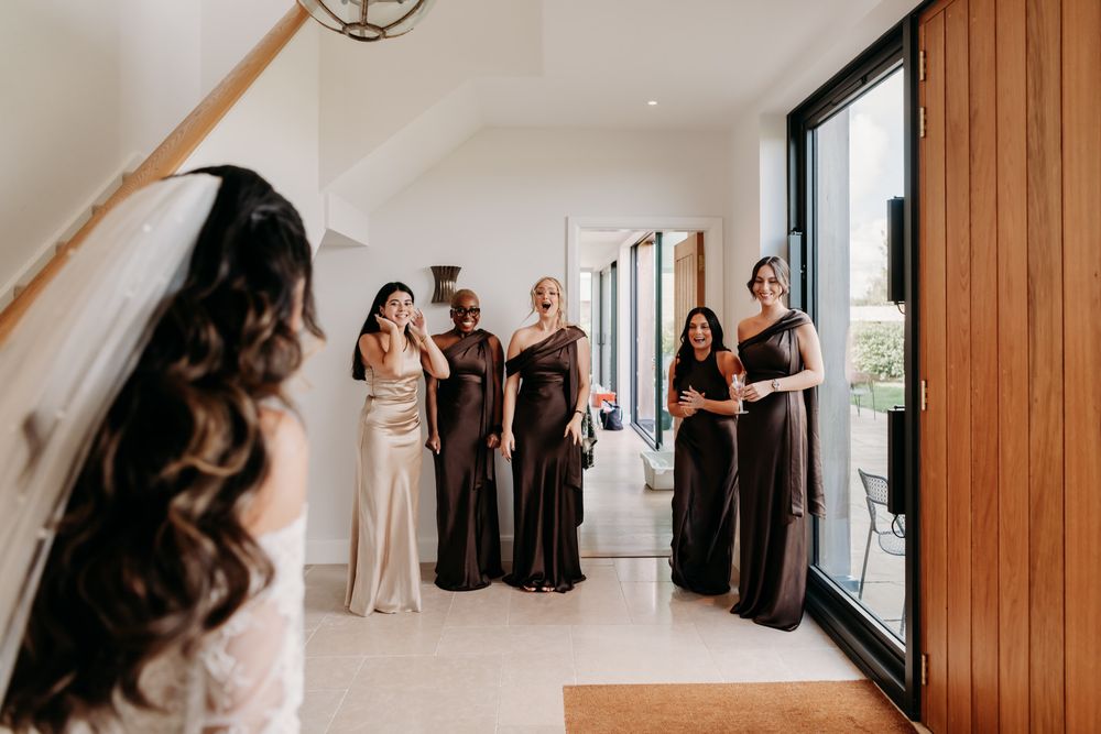 Bridesmaids in elegant black and champagne dresses gather in a bright hallway with large windows and wood paneling.