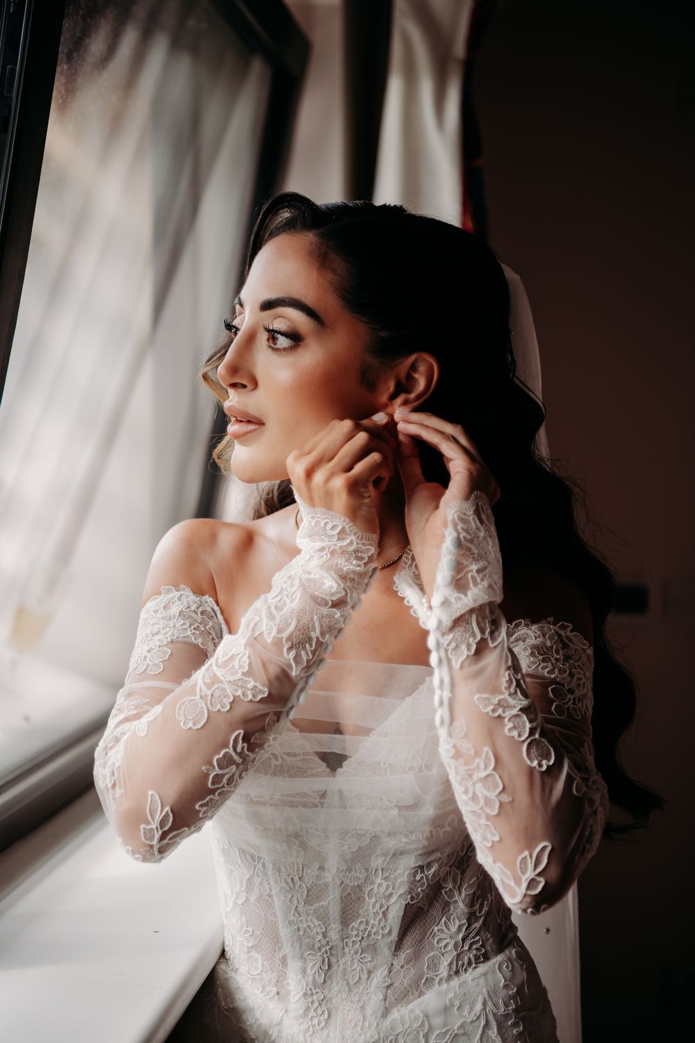 Bride in delicate lace long sleeve wedding dress gazes thoughtfully by window in romantic bridal portraits.