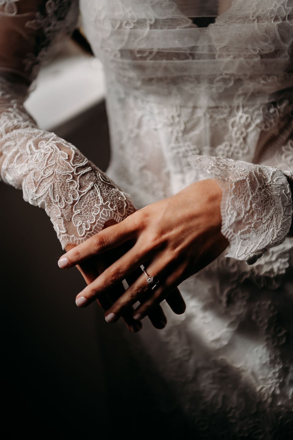 Close-up view of delicate lace sleeves and hands with an engagement ring in moody lighting.