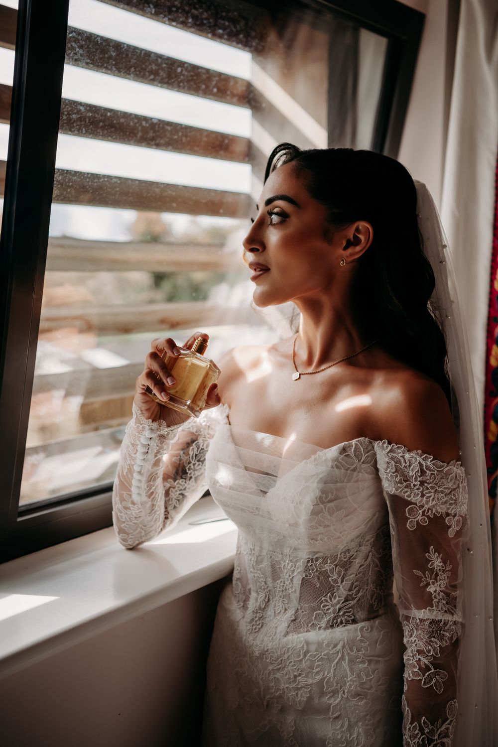 A person in a white lace wedding dress holds a cup while gazing through window blinds in warm natural light.