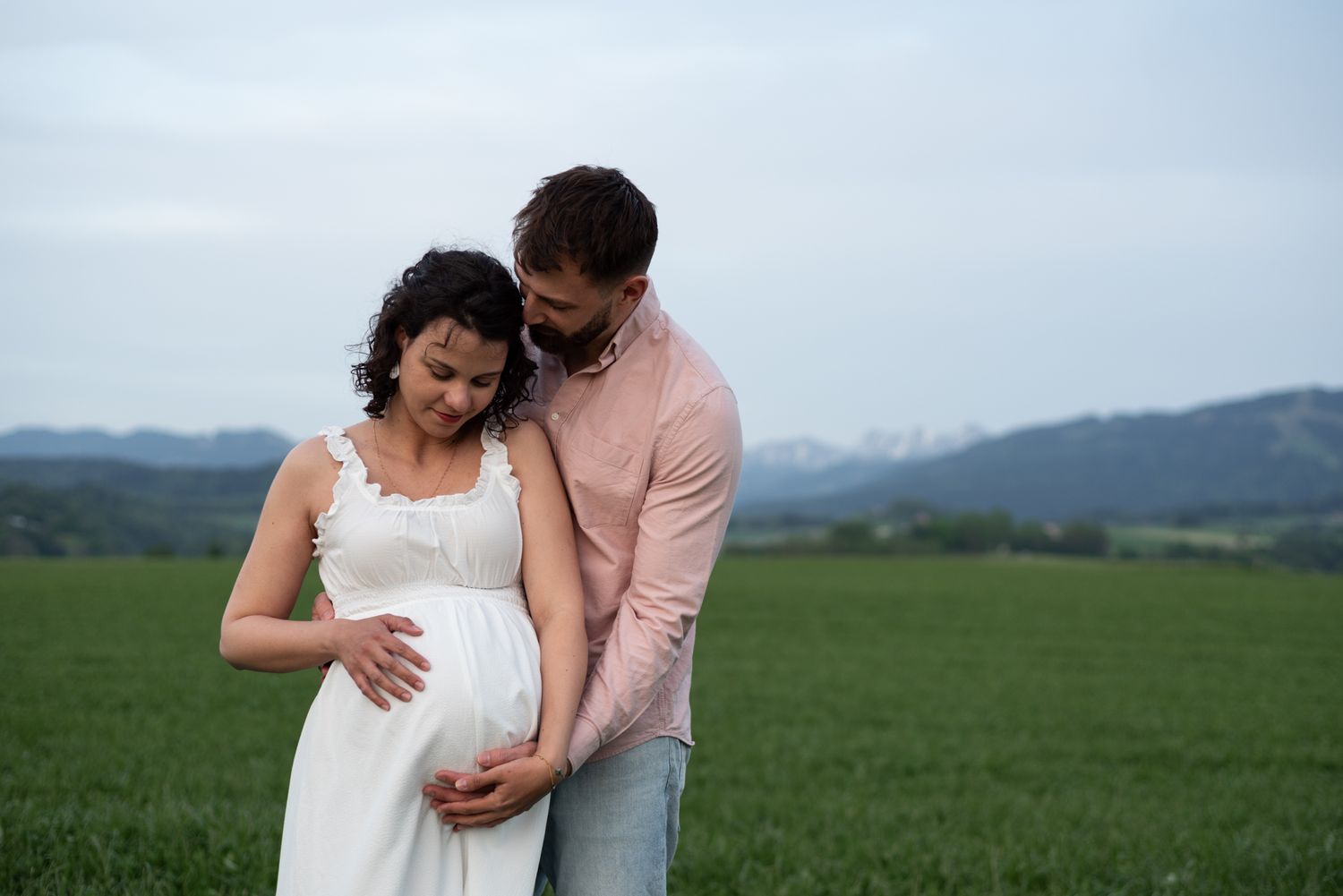 Romantic couple embracing in a green field with mountains in the background.