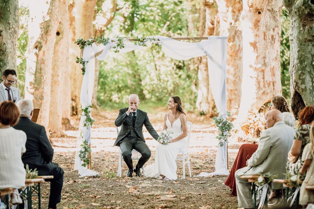 A couple embraces in a romantic outdoor setting with stone ruins and sunlit trees creating a dreamy atmosphere.