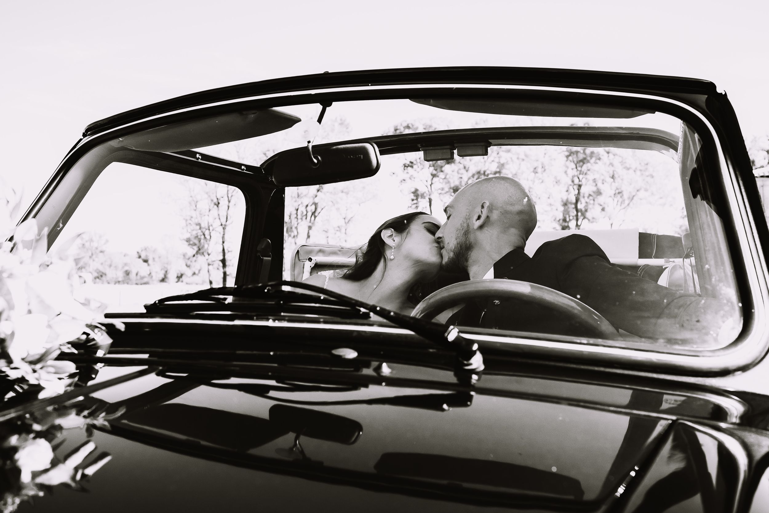 A black and white photograph of a romantic moment captured through the windshield of a vintage car.