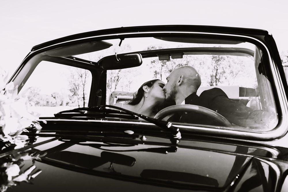 A black and white photograph of a romantic moment captured through the windshield of a vintage car.
