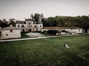 A wedding couple walks across a lush green lawn in front of a grand white historic estate building.
