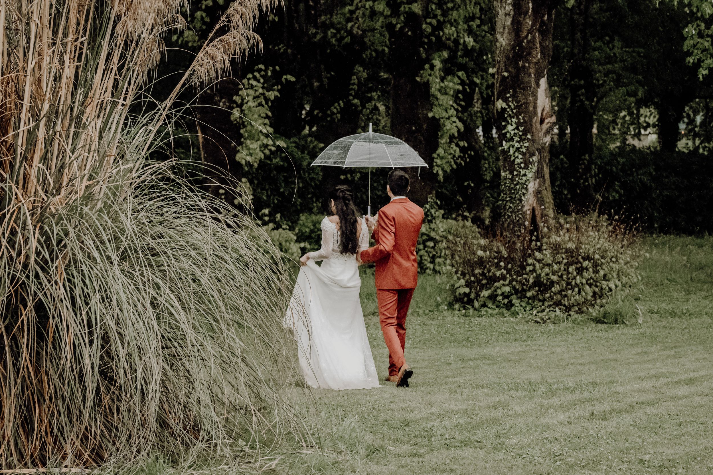 Bride in white dress and groom in orange suit share umbrella while walking through garden. photographe mariage Bordeaux, Gironde