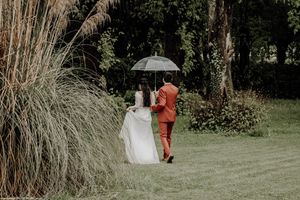 Bride in white dress and groom in orange suit share umbrella while walking through garden. photographe mariage Bordeaux, Gironde