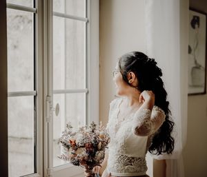 Bride with bouquet gazes out window in elegant lace wedding dress. photographe mariage Bordeaux, Gironde