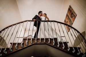 A romantic couple shares an intimate moment on a curved wooden staircase with elegant dark railings.