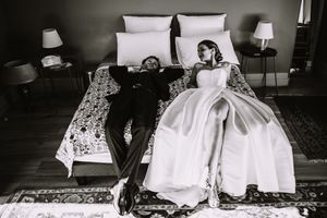 A black and white photograph shows a bride in a flowing ball gown and companion lounging on an ornate bed.