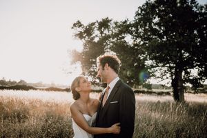 A couple in wedding attire embrace in a sunlit meadow at golden hour with trees silhouetted in the background.