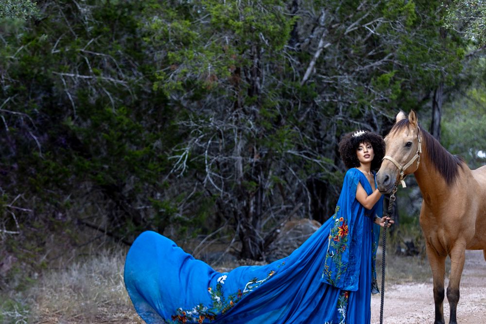 A flowing blue dress billows dramatically next to a brown horse against a natural outdoor backdrop.