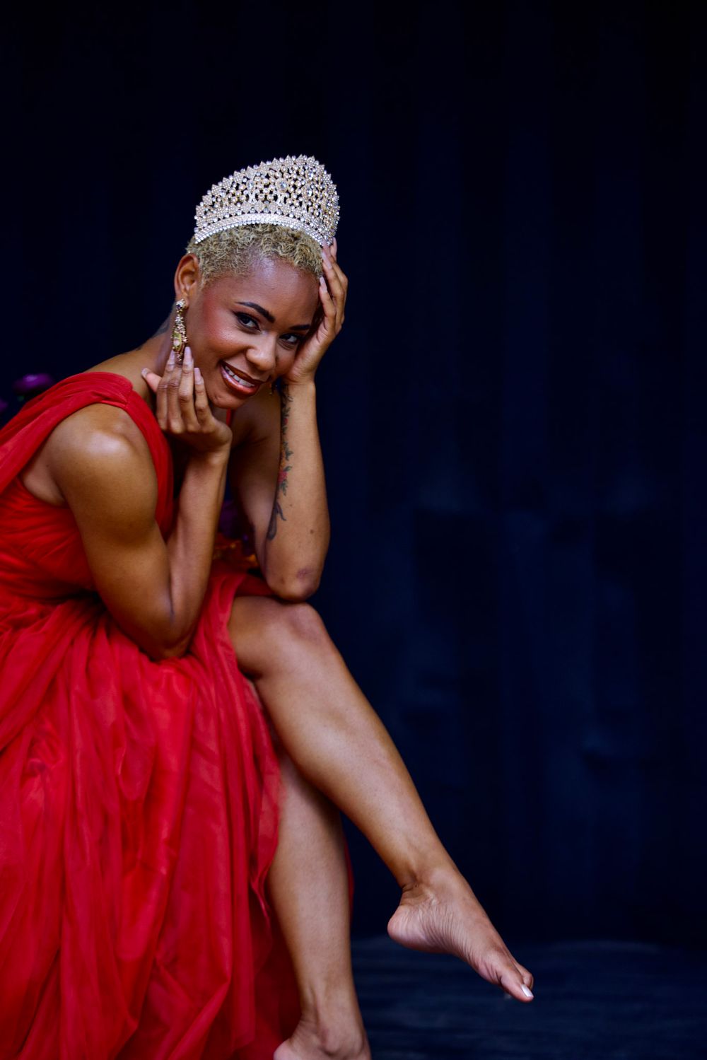 An elegant pose in a red gown and crown against a dark background with artistic lighting.