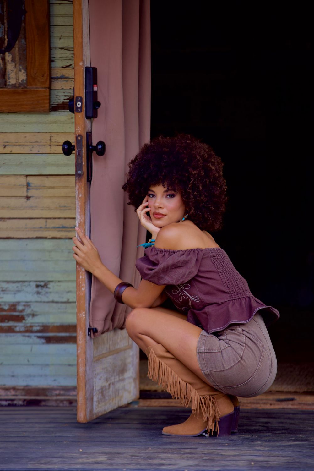 A rustic portrait by a weathered door wearing purple top and fringed boots.
