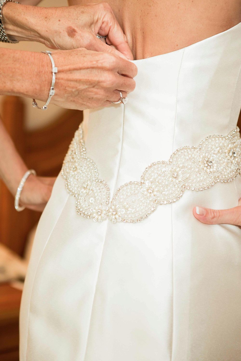 Wedding dress detail showing intricate lace and beadwork on ivory white fabric being fastened or adjusted by hands.