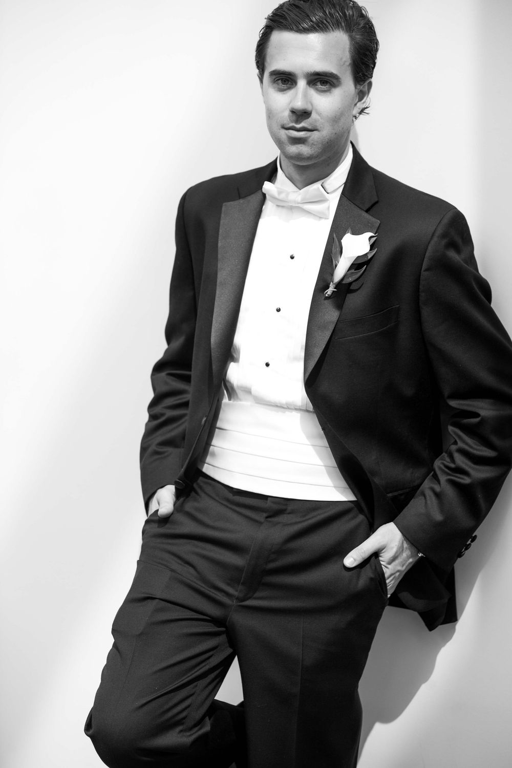 A black and white portrait of someone in a formal tuxedo with white shirt and boutonniere leaning against a wall.
