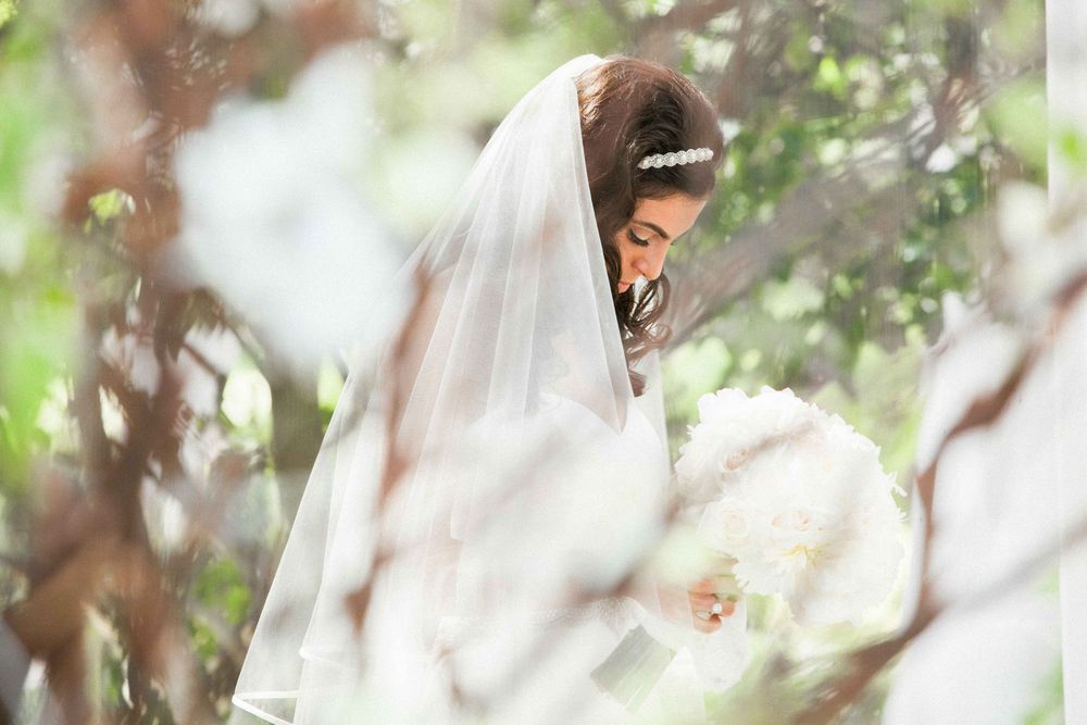 Bride in white wedding dress with flowing veil photographed through delicate bokeh in an outdoor garden setting.