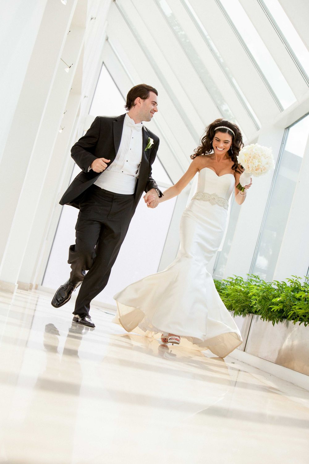 A joyful couple in wedding attire runs playfully down a bright modern hallway with white architecture and natural lighting.