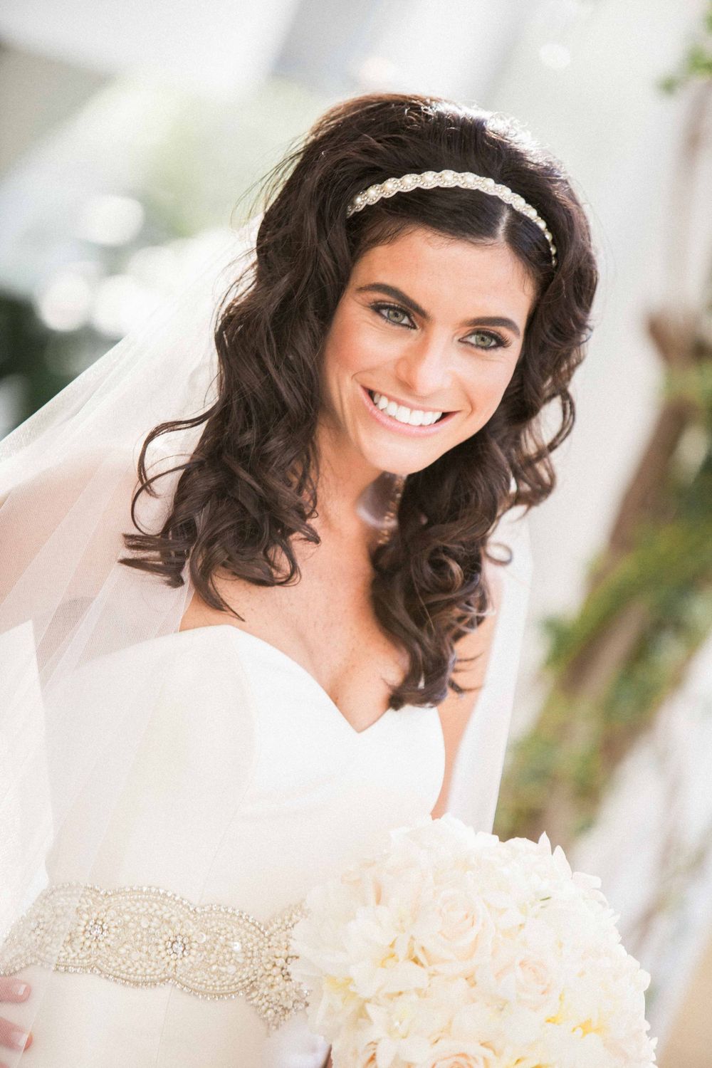 A bride in white wedding dress with dark wavy hair, beaded headband and crystal belt holds a cream colored bouquet.