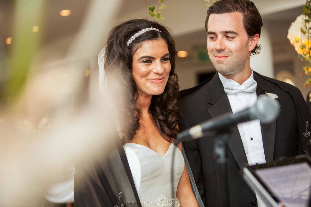 Bride and groom in formal wedding attire smile during their ceremony with soft focus foreground elements.