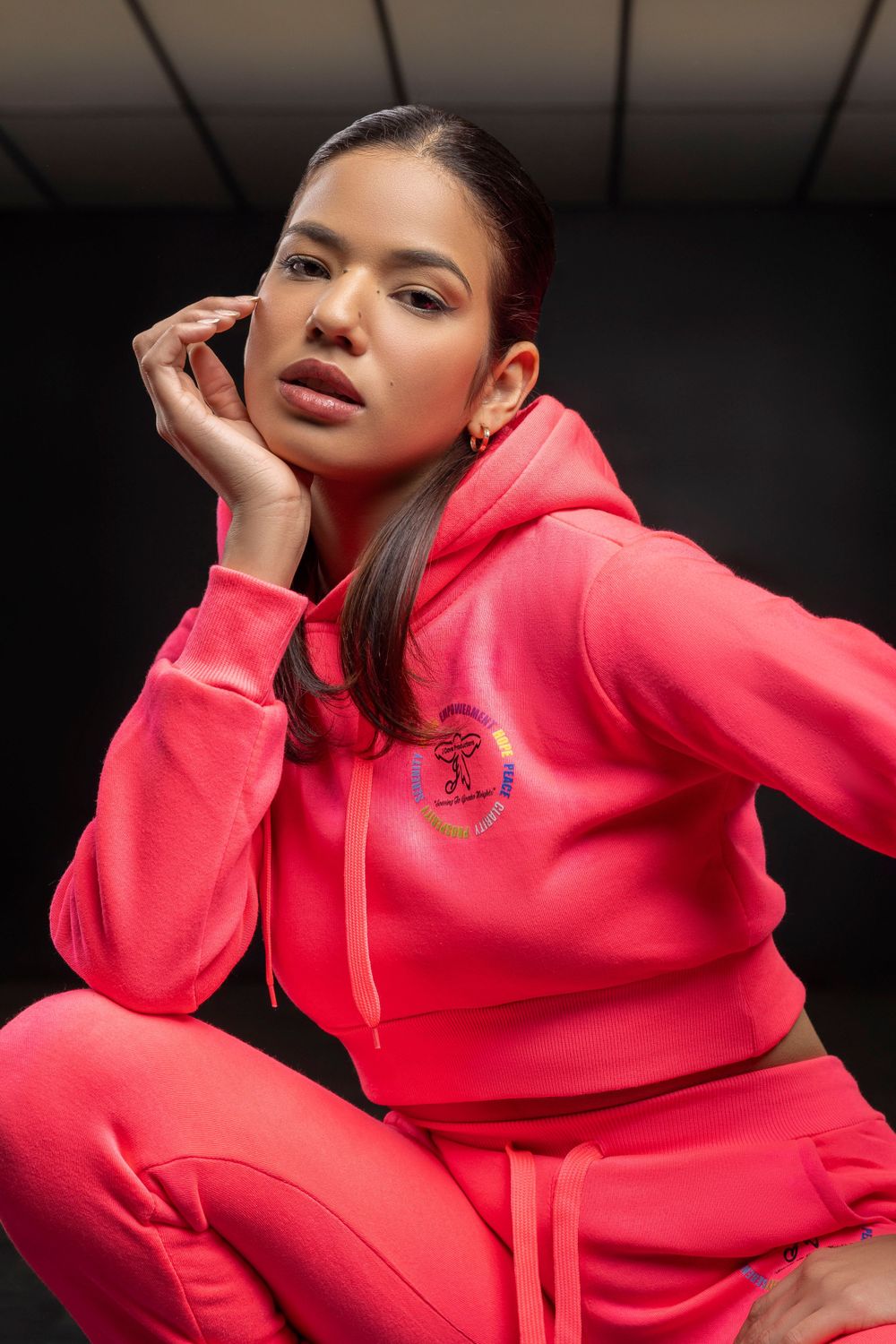 Model in bright coral pink tracksuit poses against dark studio background.