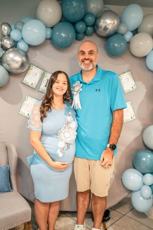 Expectant couple poses under blue and silver balloon arch at their baby shower.