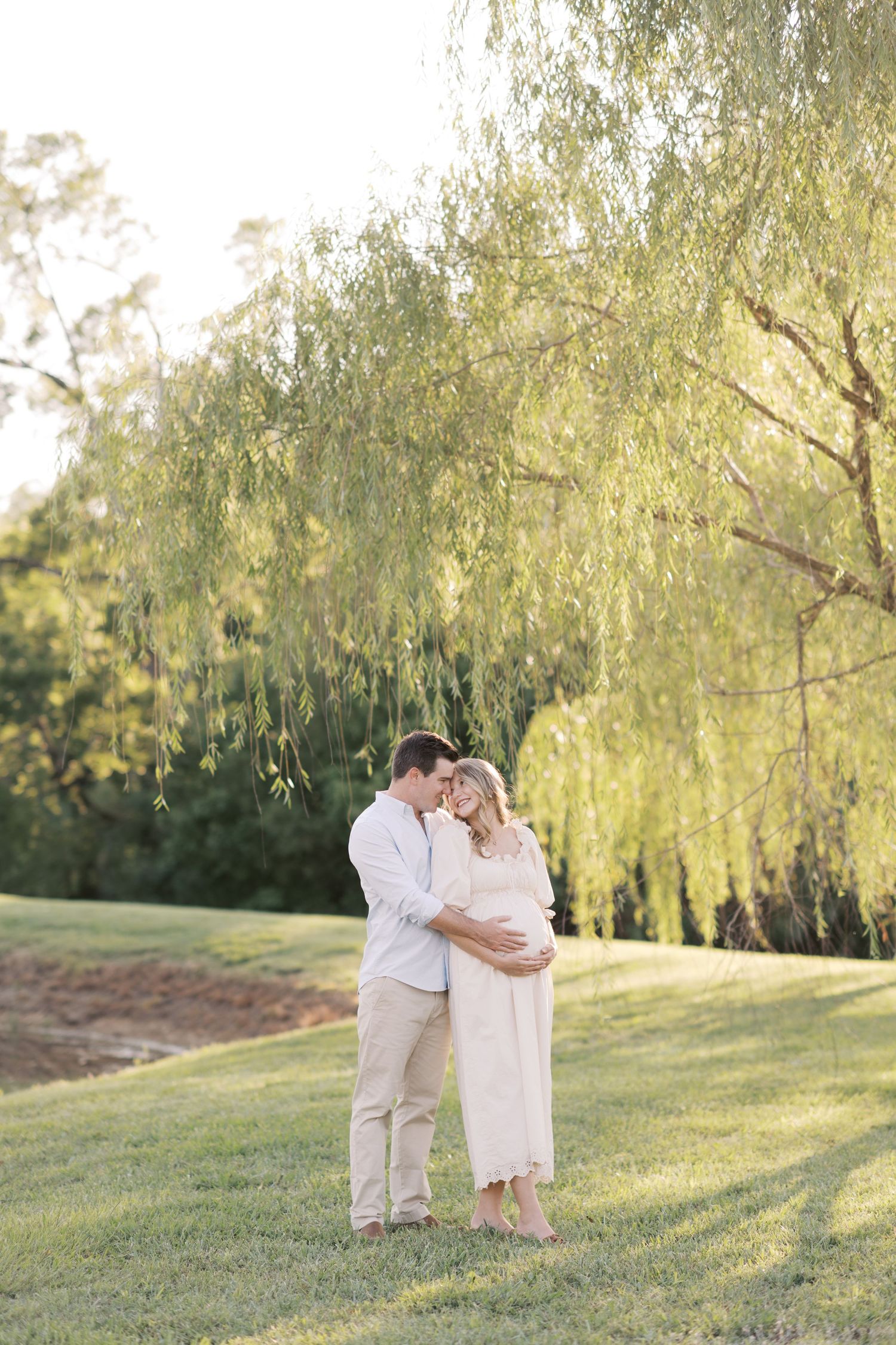 A woman in a white maxi dress holds her laughing baby under a weeping willow tree in a sunny park.