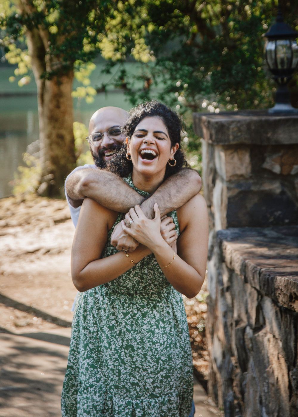 Couple shares a joyful embrace outdoors near a stone wall in natural lighting.