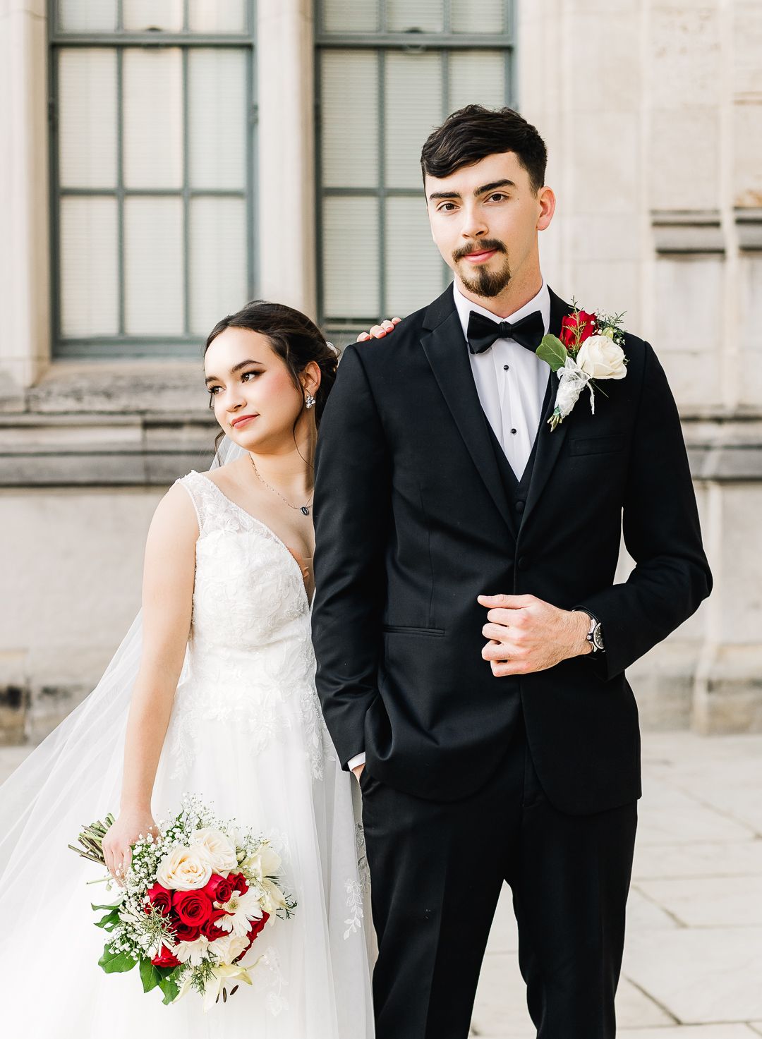 An elegant couple in formal wedding attire poses together outdoors with a red and white floral bouquet. Pittsburgh wedding Photographer