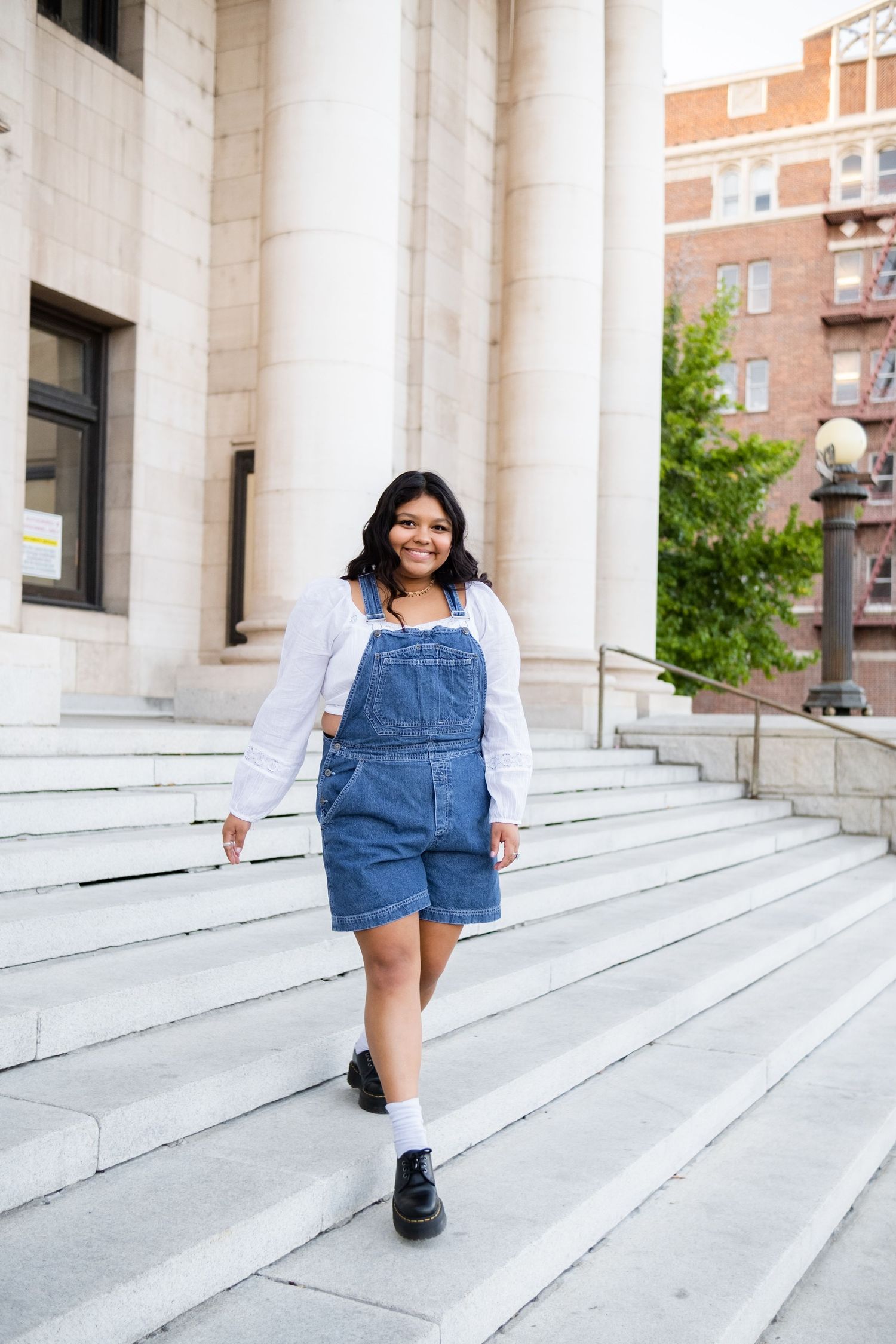 Senior in denim overall dress and white blouse walks down concrete steps near brick building downtown Reno.
