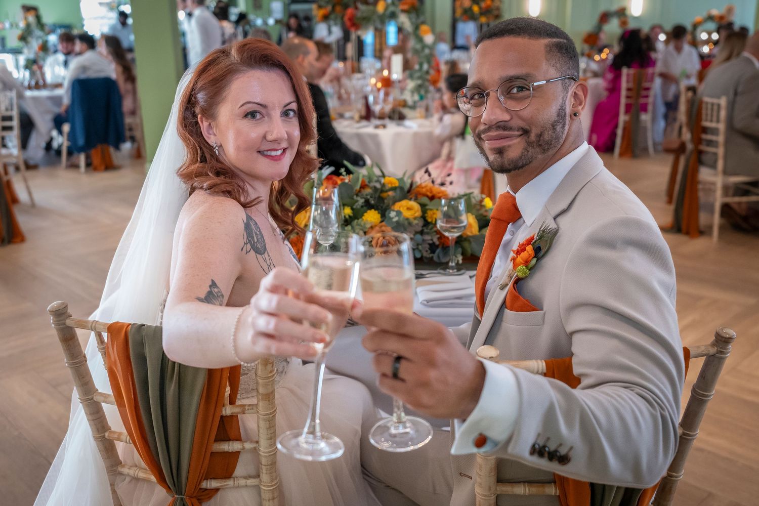 Newlyweds toast with champagne glasses at their reception featuring bright orange floral decor.