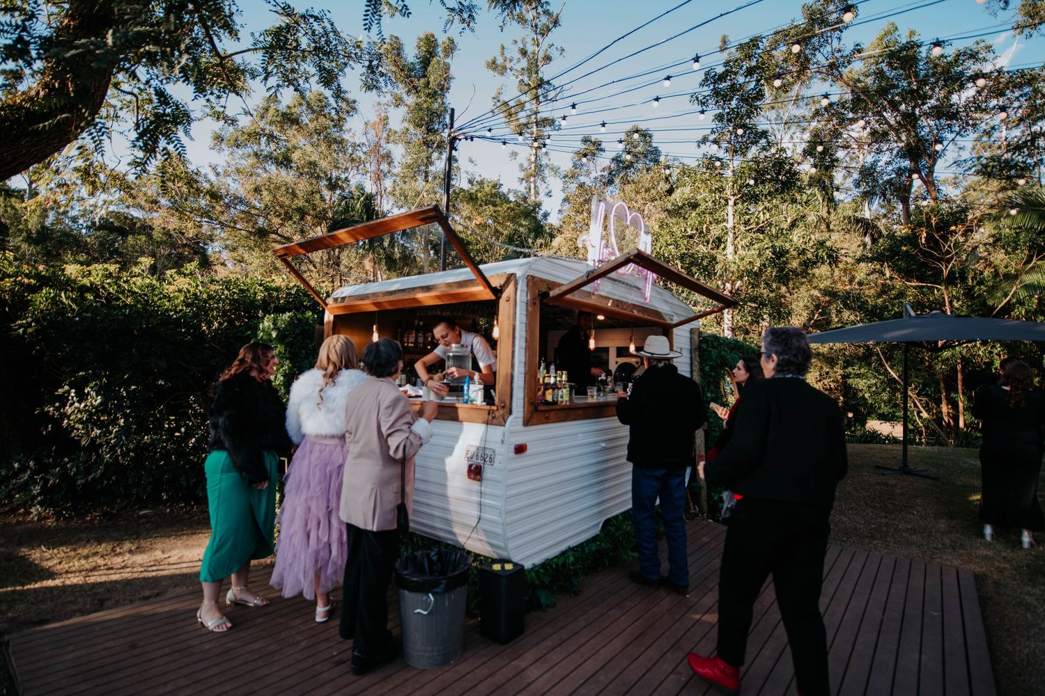 Wedding guests gather around a white food cart at an outdoor reception.