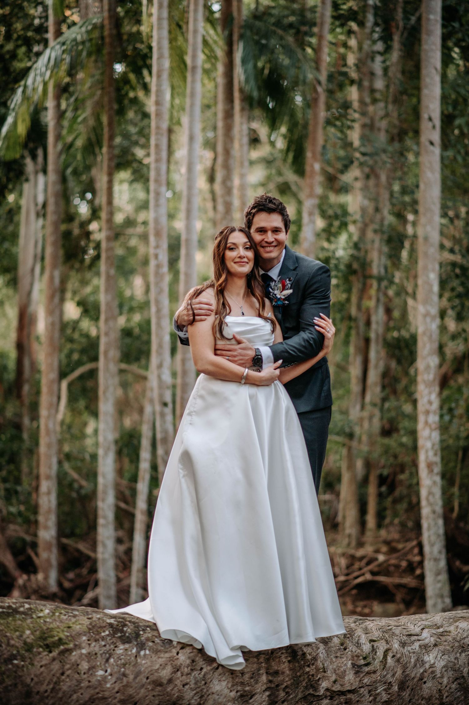 A newlywed couple poses together on a fallen tree trunk in a eucalyptus forest.