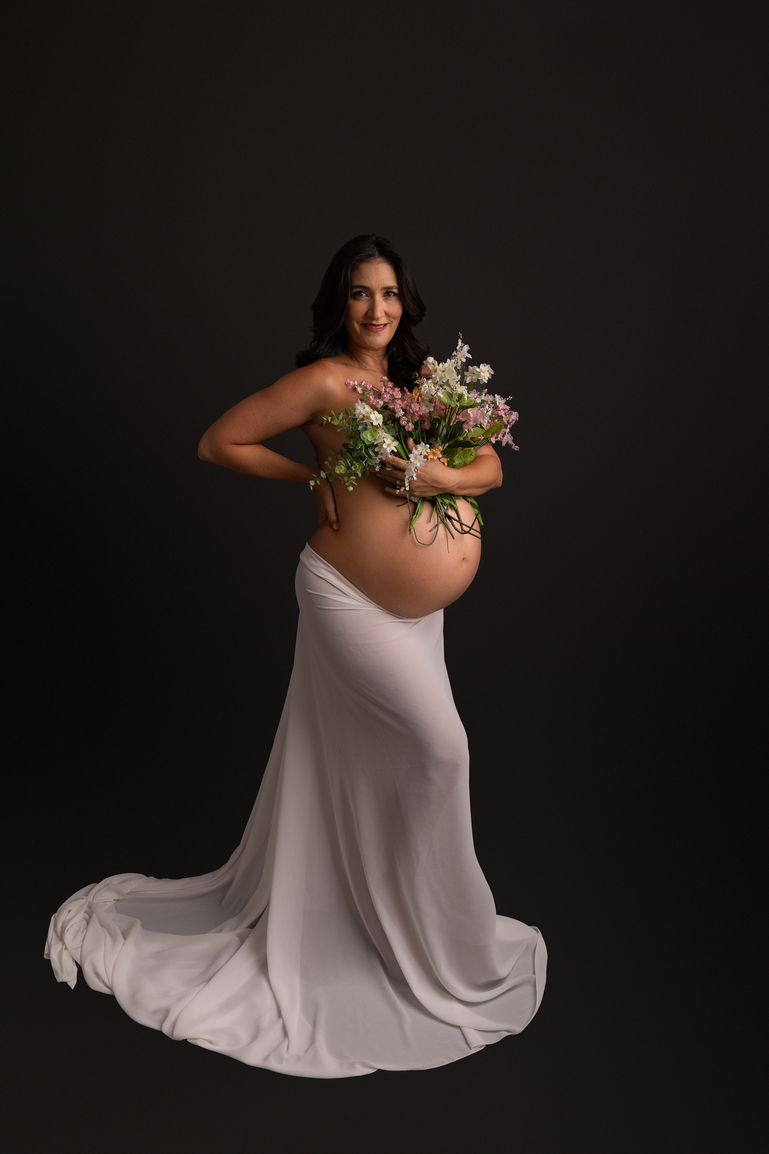 Elegant maternity woman in flowing white gown holding beautiful floral bouquet against dark background for maternity photos in Tampa