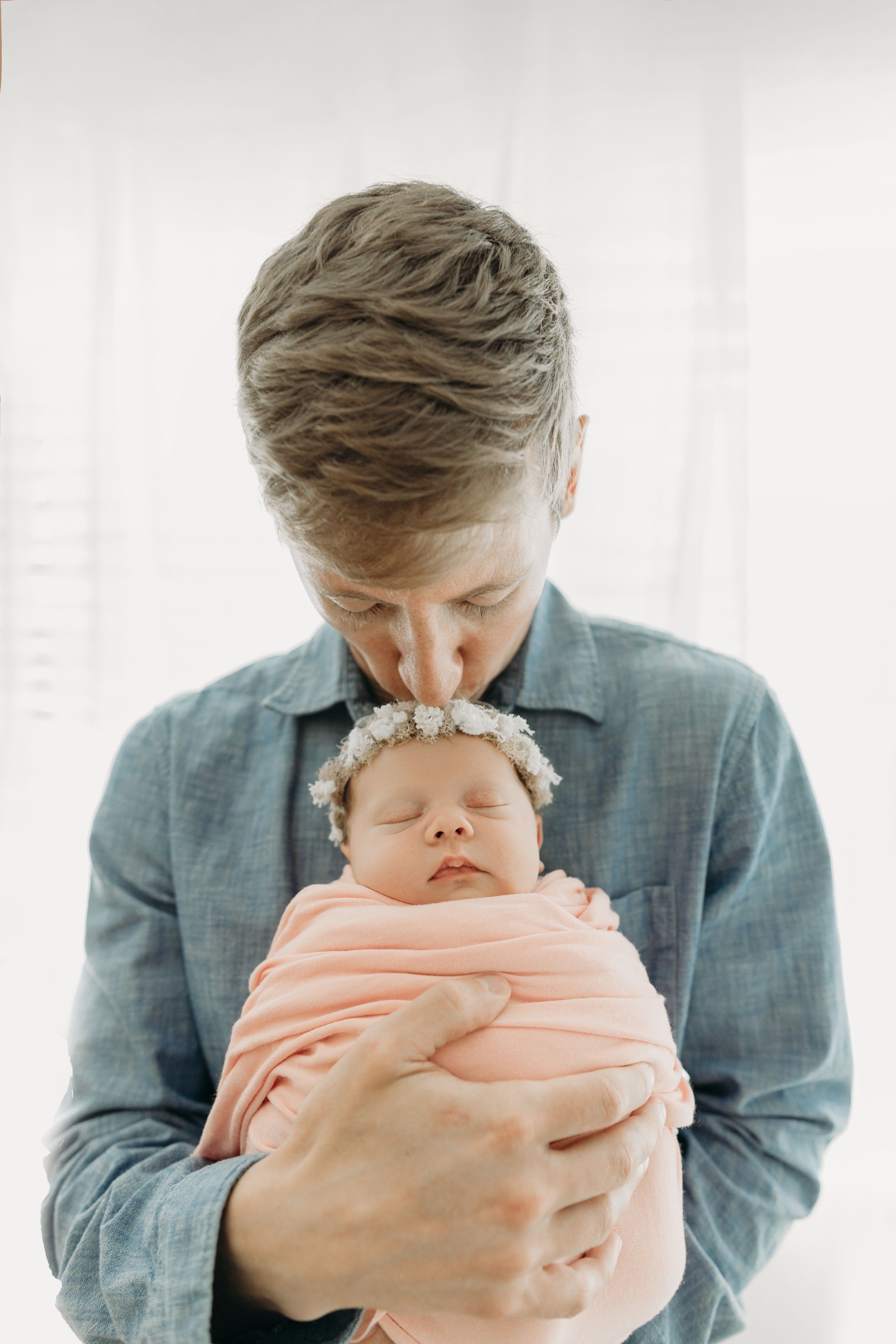 Father in a blue shirt holds and kisses sleeping newborn baby wrapped in a peach blanket during newborn photos in Tampa