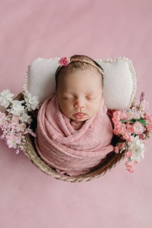 Sleeping newborn wrapped in pink swaddle rests in woven basket with floral decorations on pink backdrop, Tampa Newborn photographer