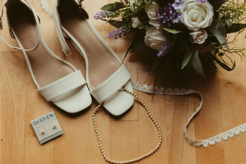 White bridal heels, pearl necklace, stud earrings, and floral bouquet wedding accessories flat lay.